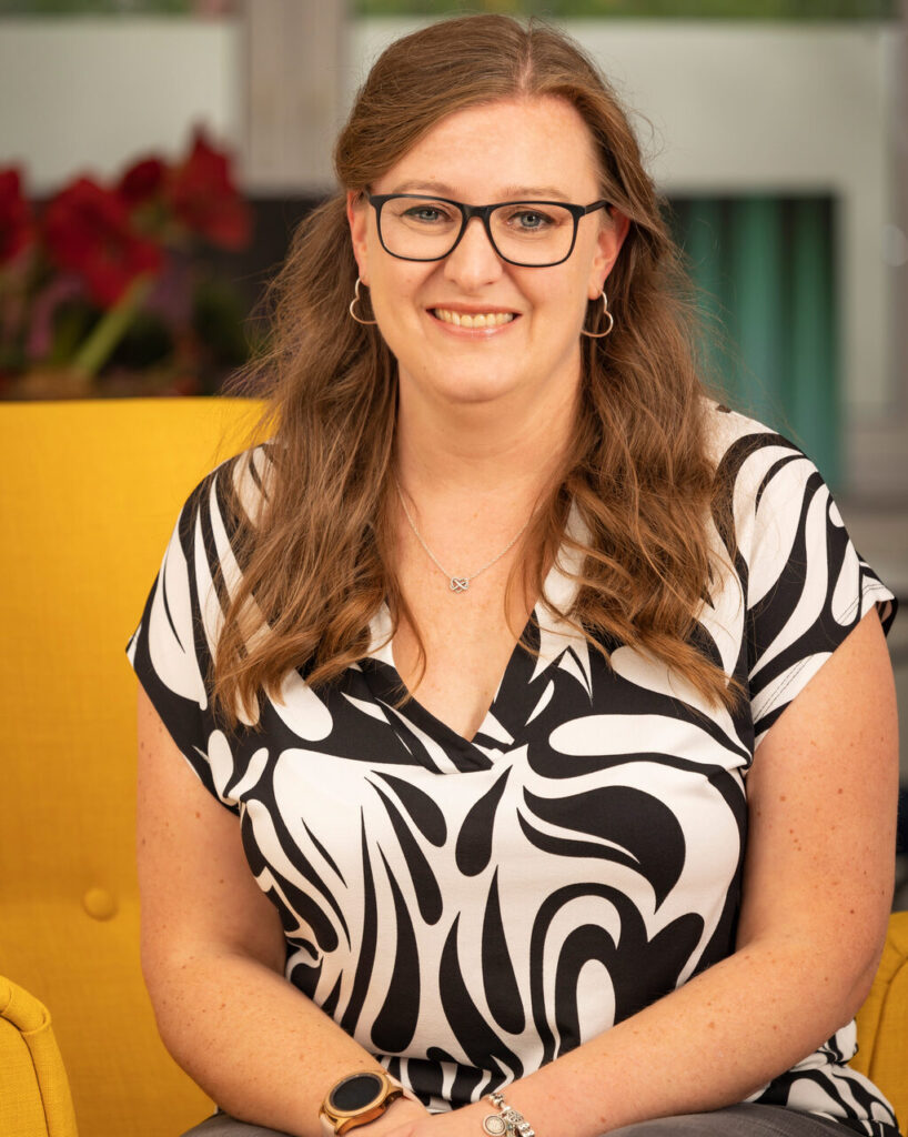 Frau mit langen, dunkelblonden Haaren, Brille und Herz-Ohrringen, schwarz-weiß gemustertes Shirt;