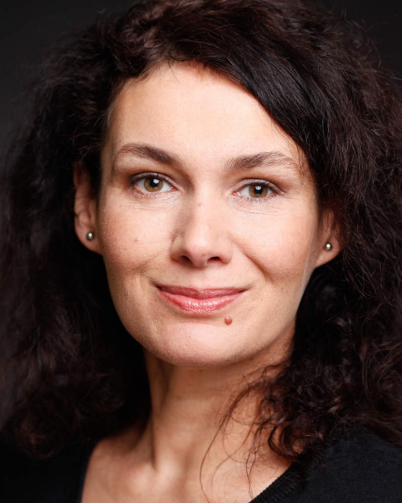 Portrait von Frau mit langen, gelockten Haaren, dunklen Augen und kleinen Ohrsteckern.