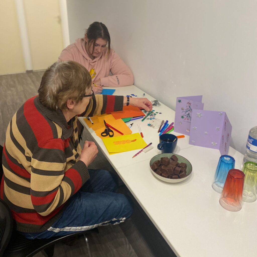 Zwei Personen sitzen am Tisch und basteln. Daneben ist eine Schale mit Dominosteinen und Gläser.