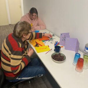 Zwei Personen sitzen am Tisch und basteln. Daneben ist eine Schale mit Dominosteinen und Gläser.