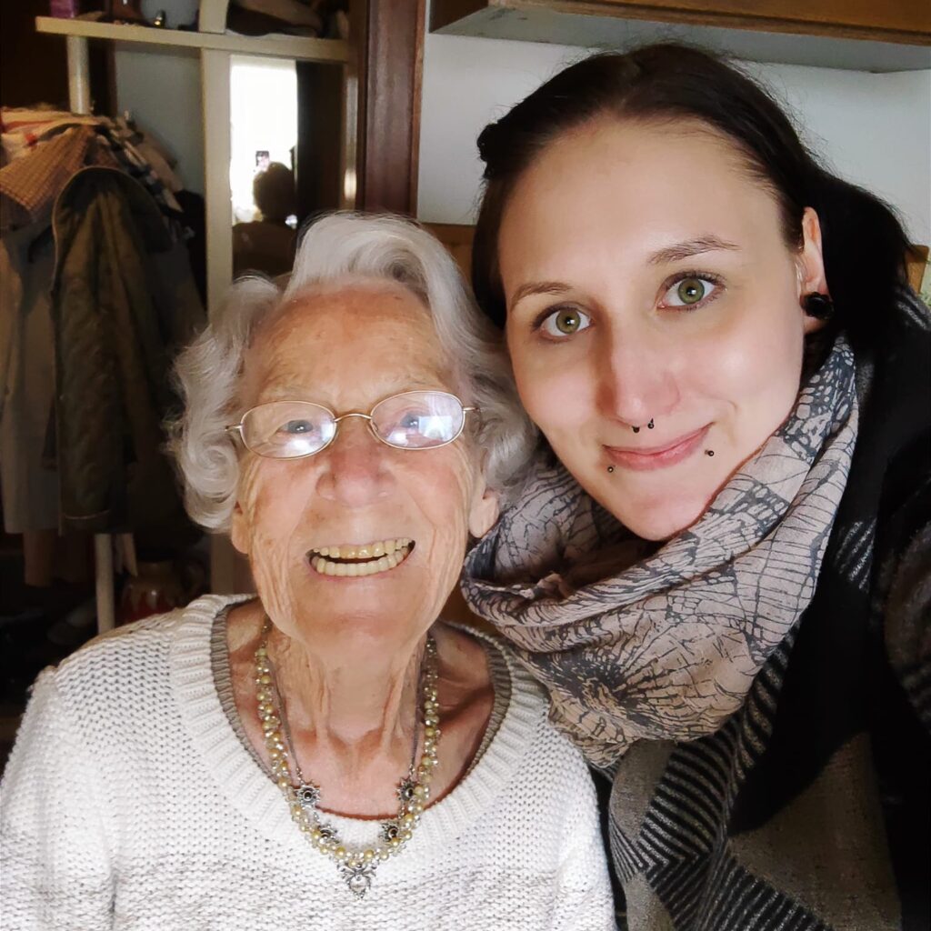 Junge Frau mit langen dunklen Haaren und ältere Frau mit grauen gewellten Haaren und Brille lächeln in die Kamera.