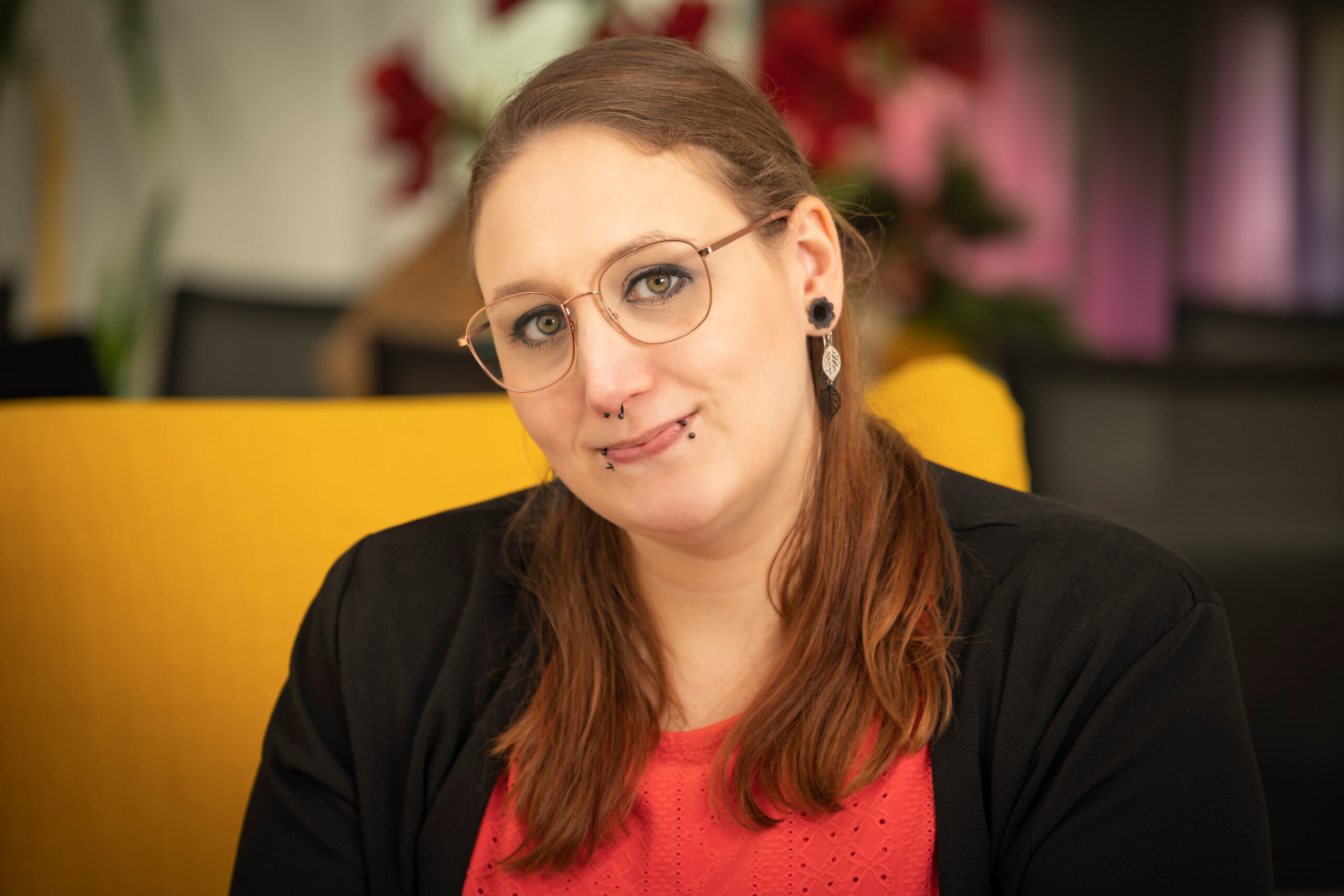 Frau mit Brille, Piercings und langem, braunen Zopf schaut in die Kamera