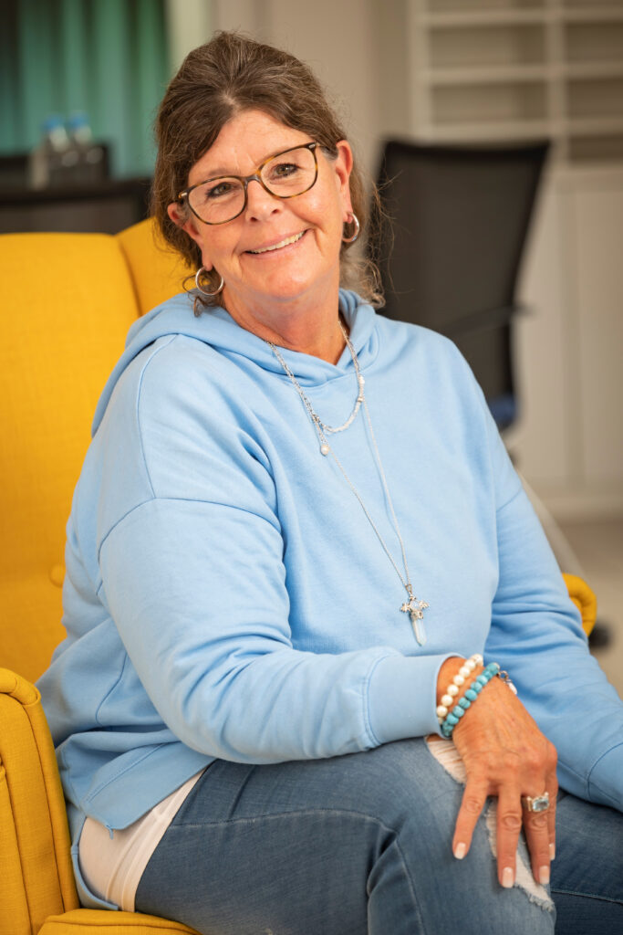 Frau mit Brille, Zopf, Schmuck und hellblauem Pullover sitzt auf dem gelben Sessel und lächelt in die Kamera