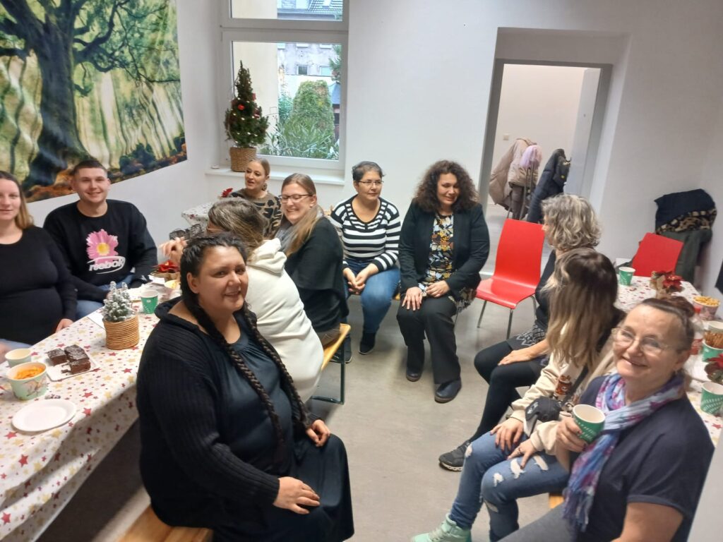 Blick von schräg oben: 11 Personen sitzen auf Bierbänken und Stühlen in einer lockeren Runde im Raum