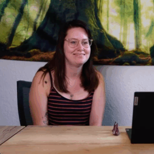 Junge Frau in gestreiftem Top, langen dunklen Haaren und Brille lächelt am Tisch