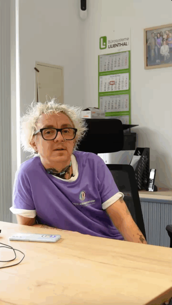 Mann mit blonden Haaren, Brille, Tattoos und Piercing in einem lila Shirt mit Logo sitzt am Schreibtisch im Büro