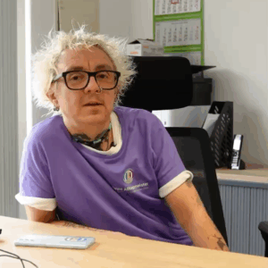 Mann mit blonden Haaren, Brille, Tattoos und Piercing in einem lila Shirt mit Logo sitzt am Schreibtisch im Büro