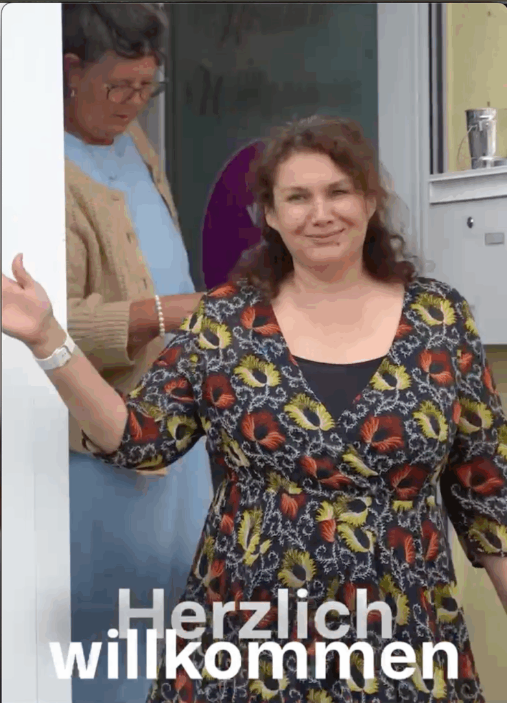 Frau mit langen dunklen Haaren, einem gemusterten Oberteil winkt in die Kamera, während sie aus der Tür geht.