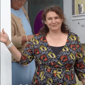 Frau mit langen dunklen Haaren, einem gemusterten Oberteil winkt in die Kamera, während sie aus der Tür geht.