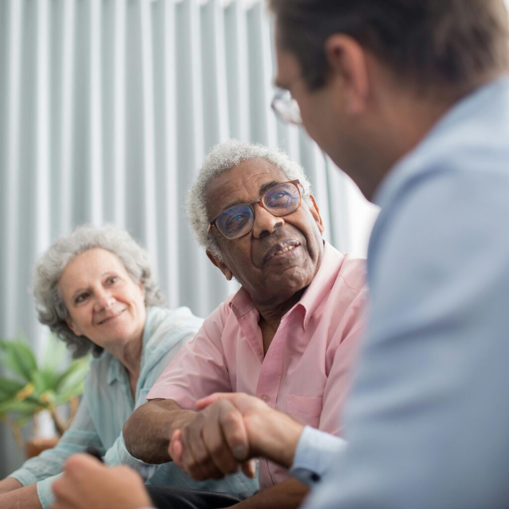 Älterer Mann schüttelt die Hand eines jüngeren Mannes. Eine Frau sitzt neben dem älteren Mann und schaut lächelnd auf das Geschehen.