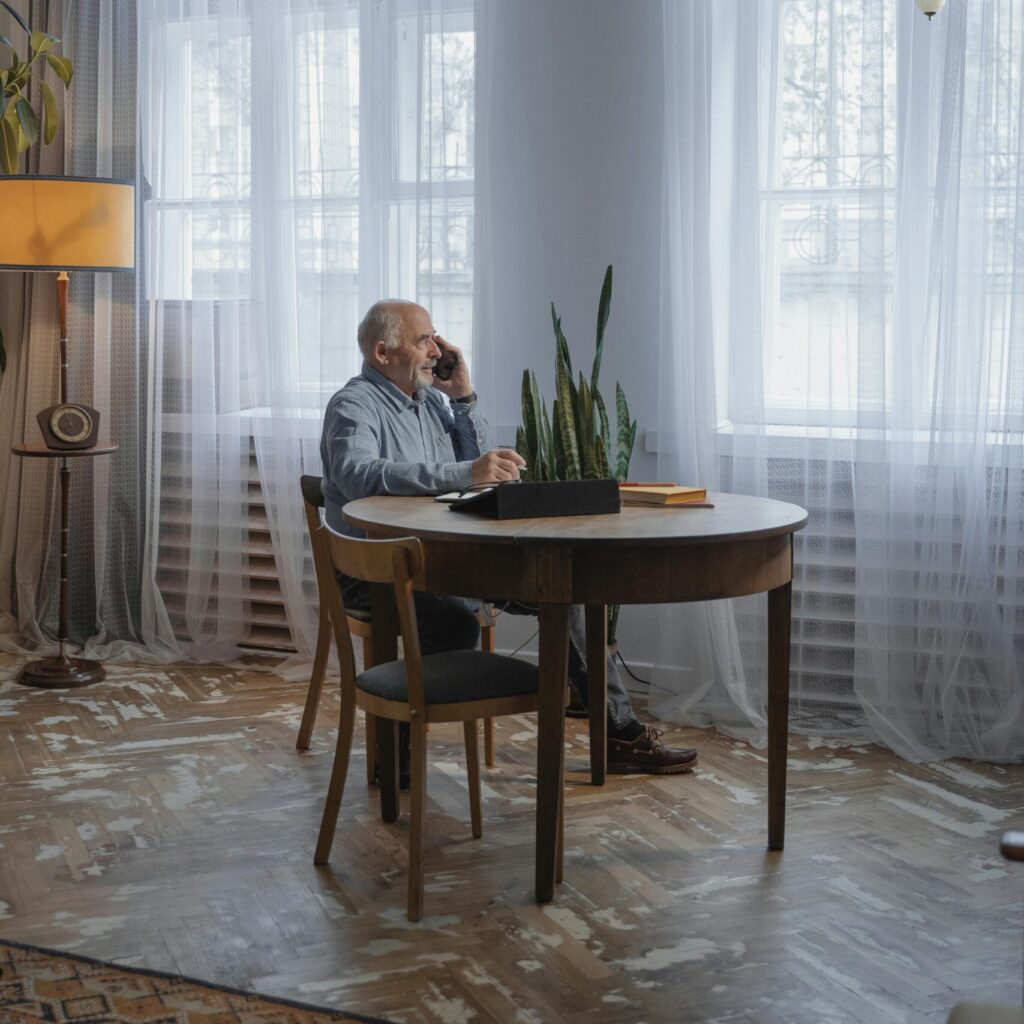 Älterer Mann sitzt am Tisch und telefoniert. Der Boden ist vintage, lange weiße Gardinen hängen vor den Fenstern.
