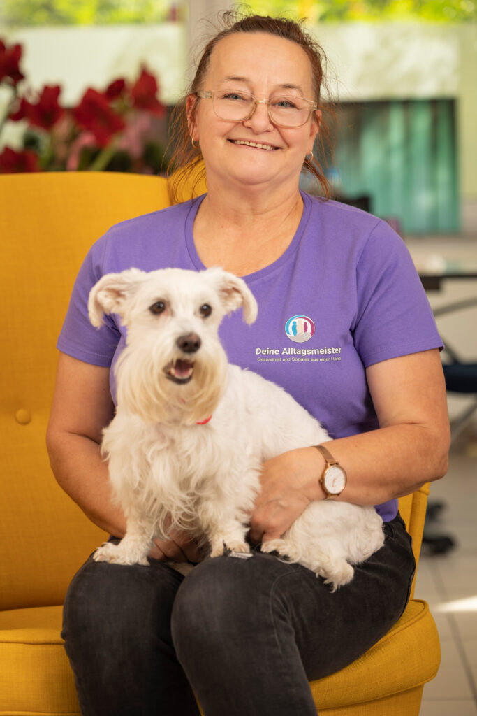 Eine Frau in lila Firmenshirt sitzt auf dem gelben Sessel, hat einen kleinen weißen Hund auf dem Schoß und lächelt in die Kamera