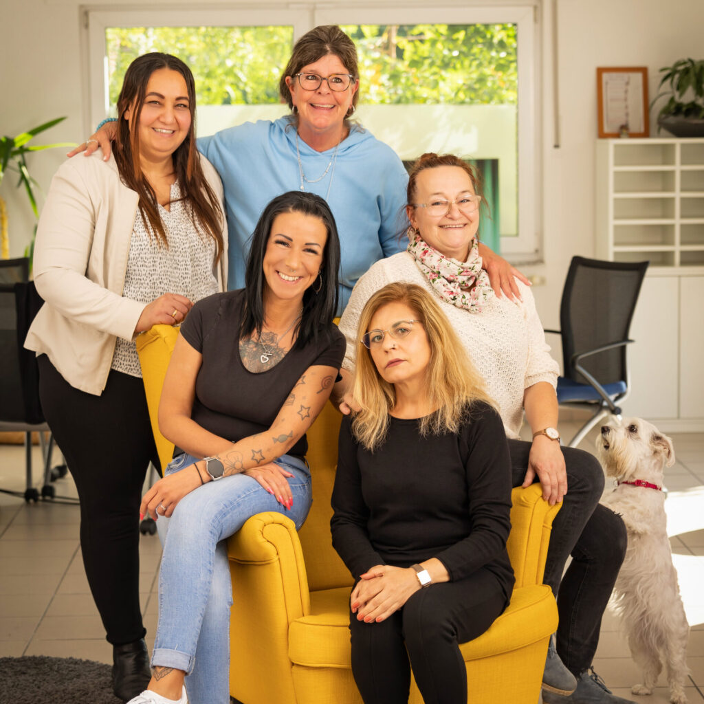 Ein kleiner Hund und fünf Personen stehen um bzw. sitzen auf einem gelben Sessel.