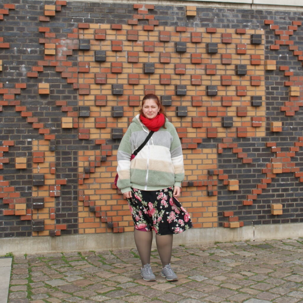 Frau in herbstlicher Kleidung steht von einer markanten Wand, wo mit roten, gelben und schwarzen Ziegelsteinen ein Muster erstellt wurde