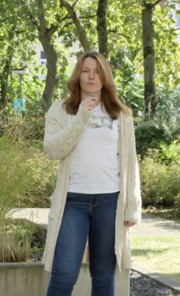 Frau mit langen Haaren steht im Wald und hält ein Mikro.